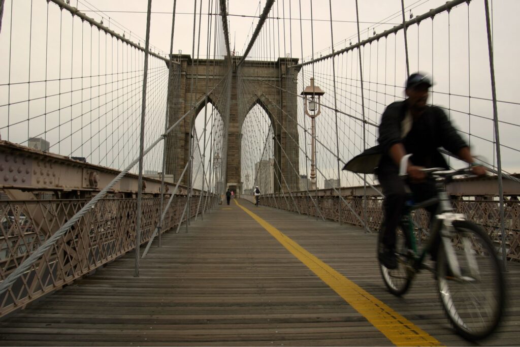 Pedestrian and cyclist on the wooden walkway of the Brooklyn Bridge, with steel suspension cables and stone towers in the distance.