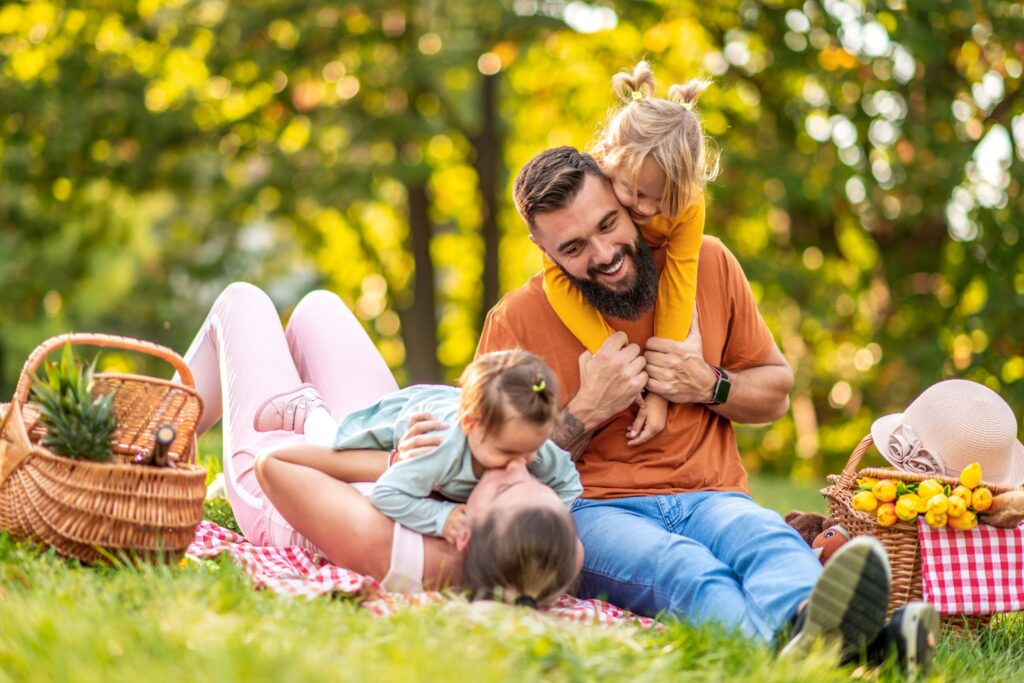 Father with a beard shares a playful picnic moment with two kids on a sunny park day, baskets of fruit nearby on a checkered blanket.