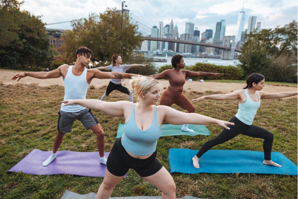 Group of people doing yoga in a park with a city skyline and a bridge in the background.