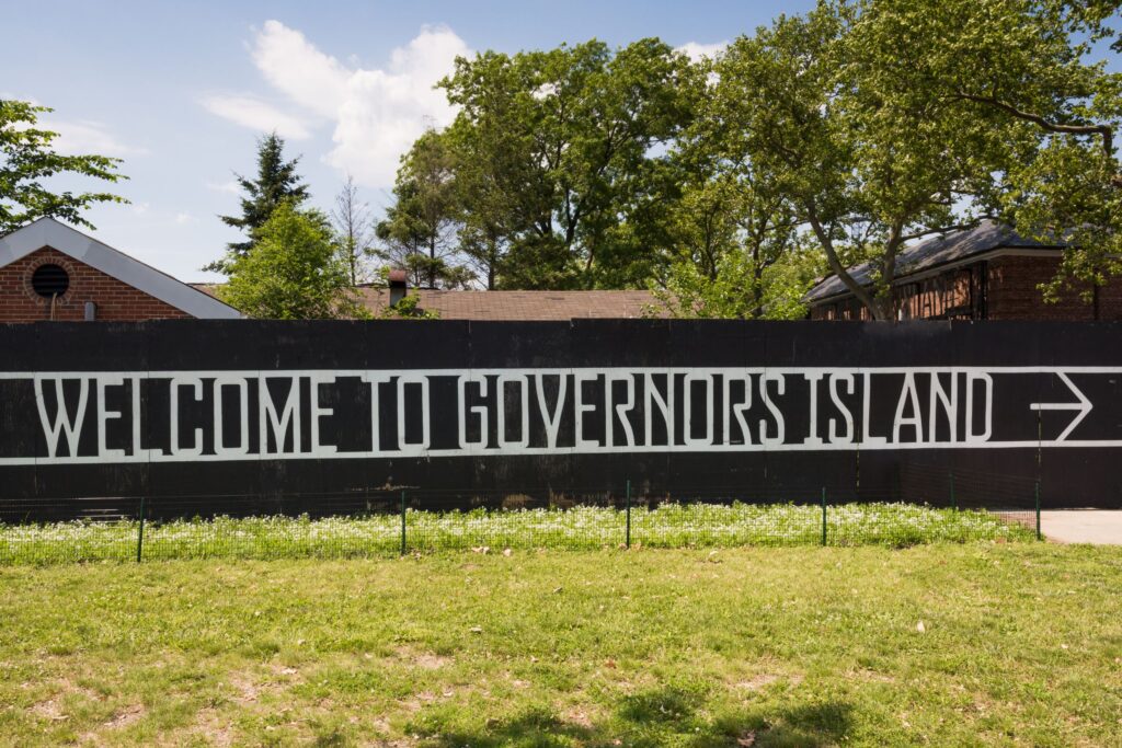 Black wall mural with large white lettering reading 'WELCOME TO ... ISLAND' beneath a blue sky with green trees and brick buildings behind it.