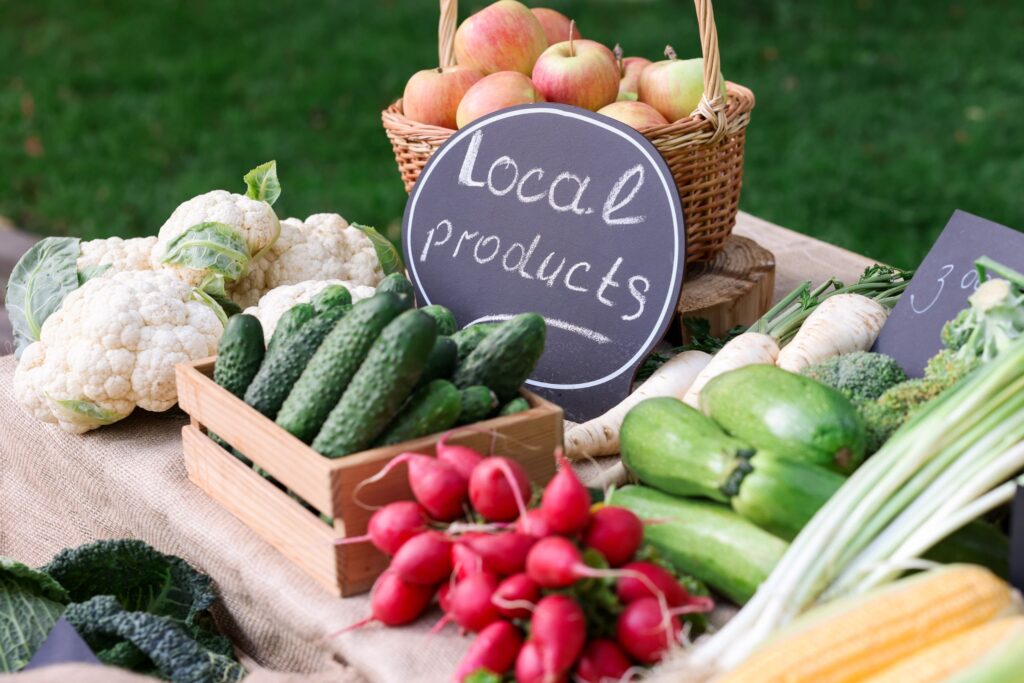 Basket of apples with a chalkboard that reads Local Produce, surrounded by cauliflower, cucumbers, and radishes at an outdoor market stall.