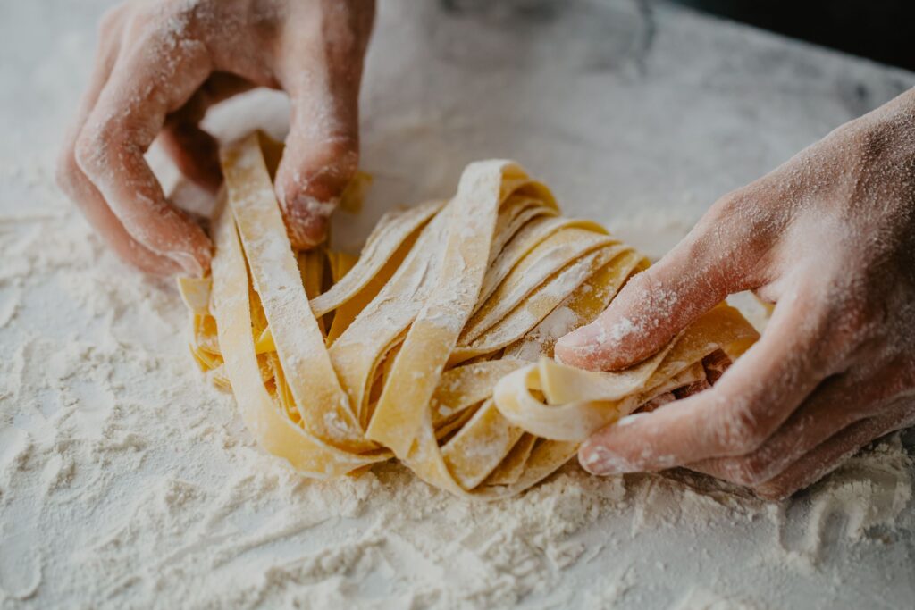 Close-up of flour-dusted hands shaping long ribbons of fresh pasta on a floury work surface.