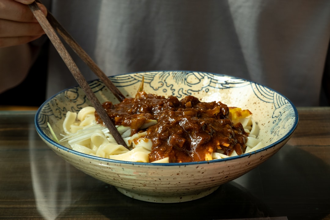 Una persona che dispone condimenti vegetali colorati su una ciotola di noodle freddi ghiacciati su un tavolo da pranzo