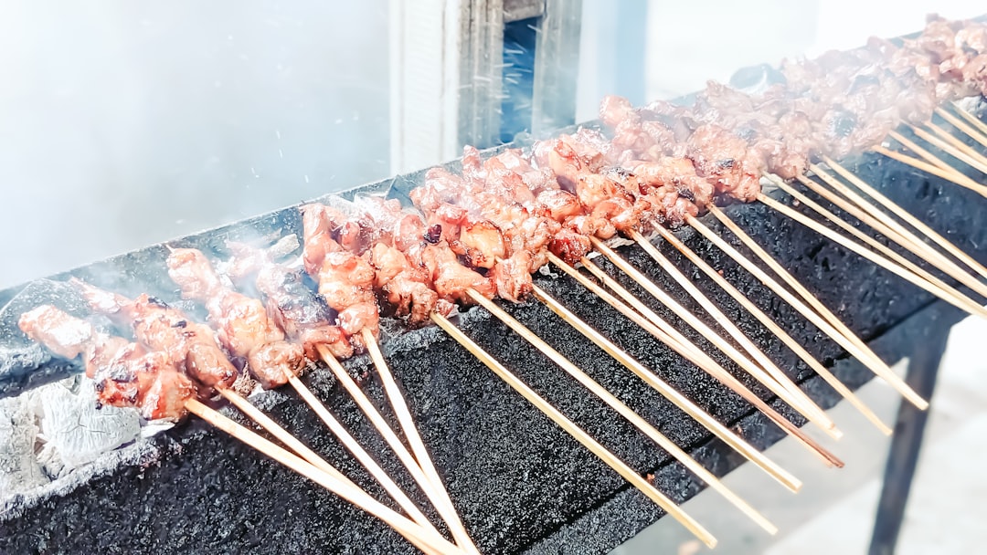 Um vendedor grelhando espetinhos de satay de comida de rua sobre carvão em uma barraca de mercado ao ar livre, com fumaça subindo ao redor da carne.