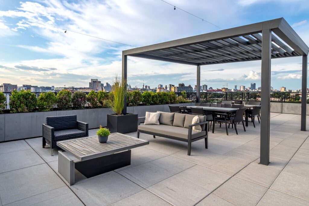 Rooftop terrace with modern gray seating, a pergola, potted plants, and a distant city skyline under a blue sky.