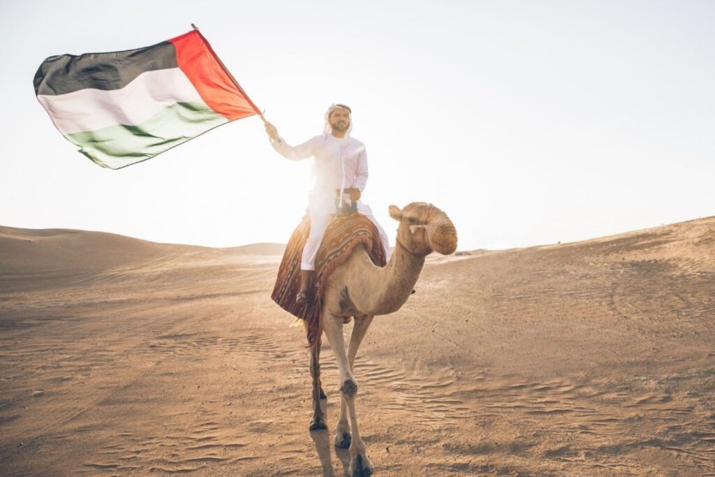 Emirati man in traditional kandura riding camel while proudly holding UAE flag during national day events, representing authentic cultural heritage celebrations in Dubai