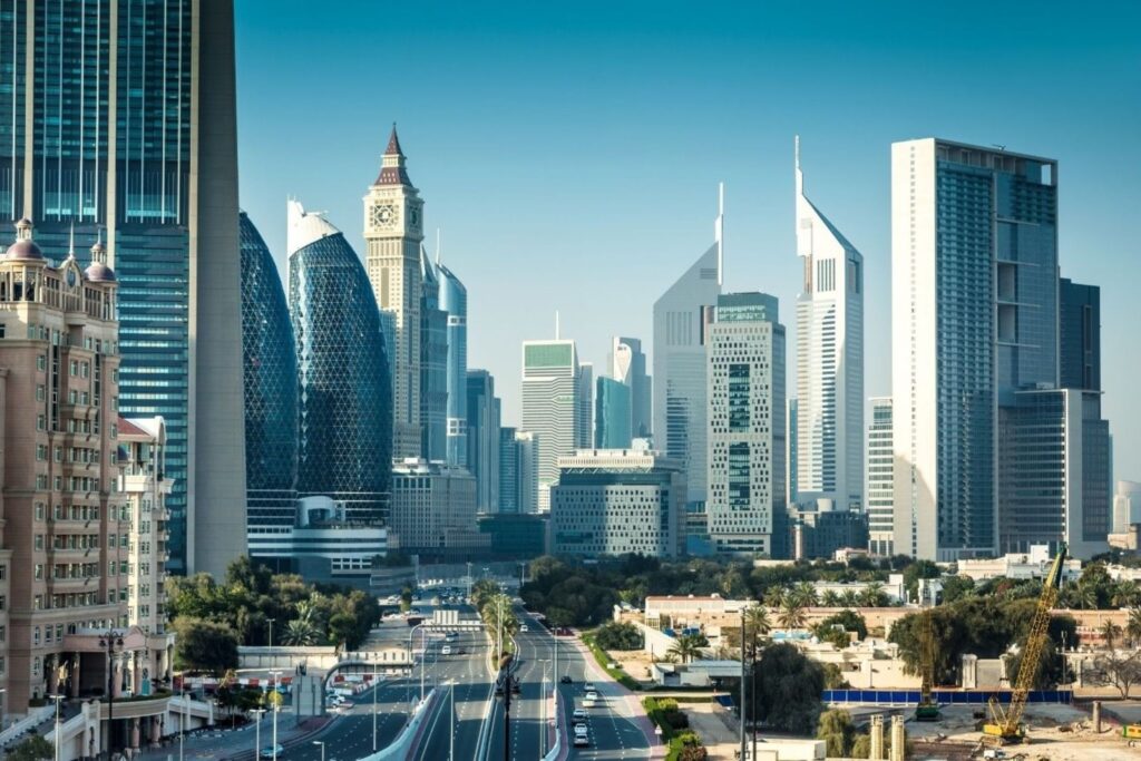 City skyline with tall modern skyscrapers and a wide multi-lane road in the foreground.