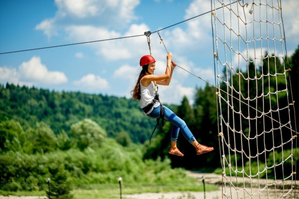 Woman wearing a red helmet and safety harness traversing a rope-net obstacle outdoors with a blue sky above.