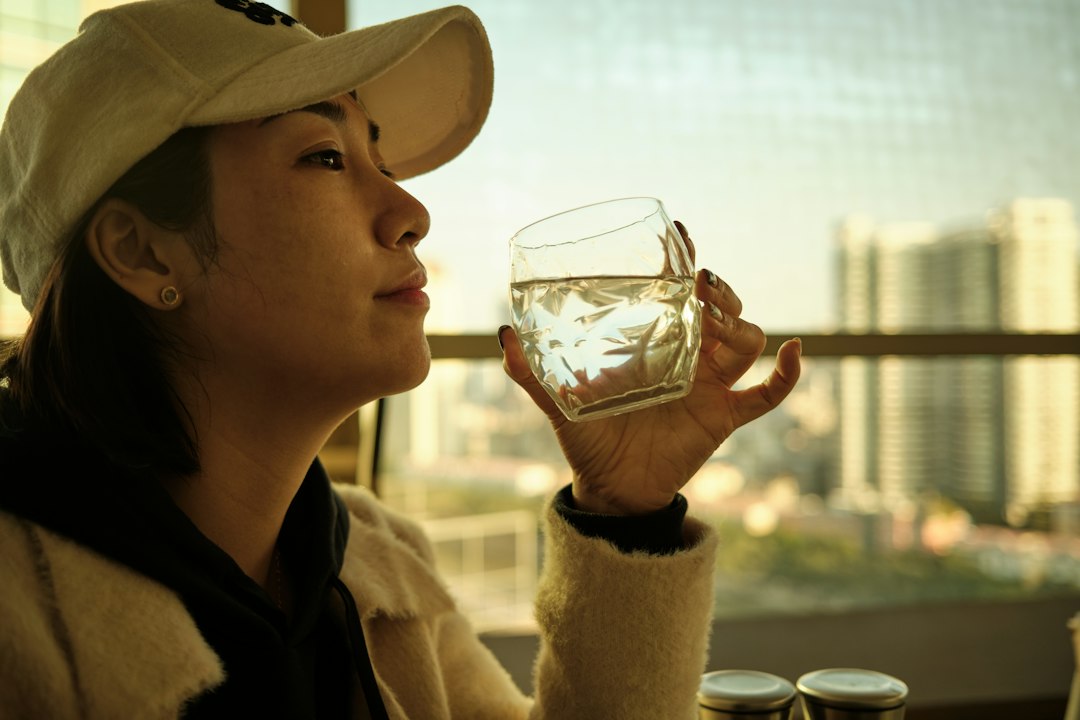 A woman drinking a glass of water by a sunlit window, supporting her Dubai skincare routine from within